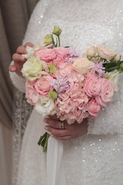 A person wearing a white, sequined wedding dress holds a round floral bouquet featuring soft pink and cream roses, pale lavender hydrangeas, and white lisianthus, with greenery accents. The bouquet is