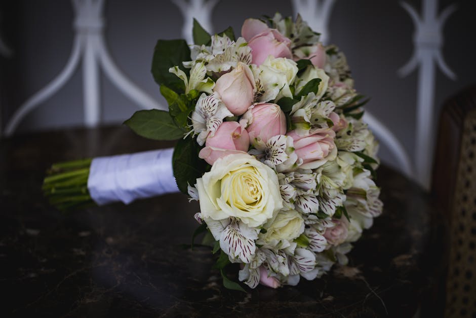 A delicate wedding bouquet featuring a mix of fresh cream and soft pink roses, complemented by white alstroemeria with dark streaks on the petals and lush green leaves. The bouquet is tightly arranged