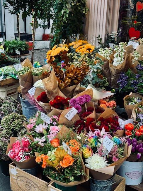A vibrant outdoor display of flower bouquets arranged in brown wrapping paper and placed in metal buckets and wooden crates. The bouquets feature a variety of fresh flowers, including bright pink lili