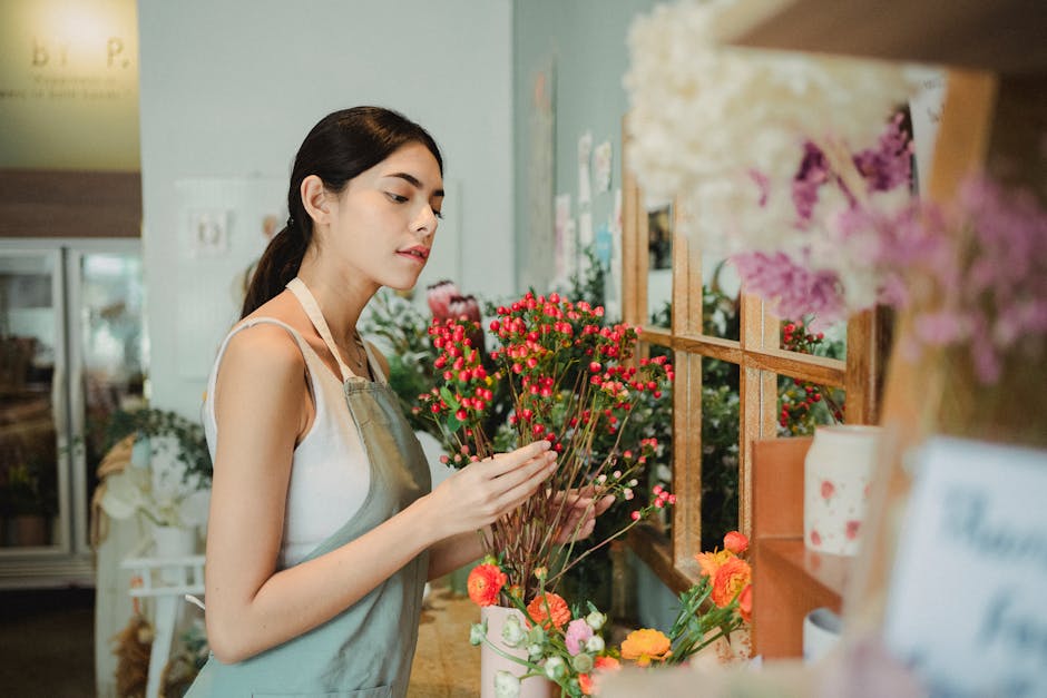 A young woman with dark hair, wearing a light-colored apron over a white sleeveless top, is examining a bouquet of fresh, vibrant pink and red flowers in a floral shop. She is holding the bouquet with