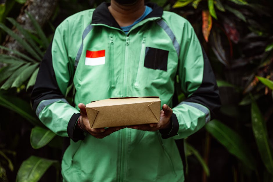 A delivery person wearing a green jacket with black and gray accents, holding a rectangular cardboard parcel with both hands. The jacket features a black patch on the chest and a small red and white f