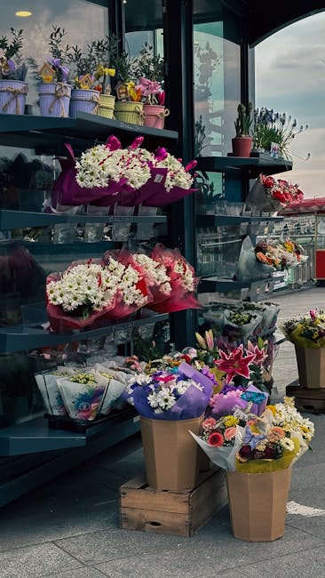 A variety of fresh flower arrangements displayed on tiered shelves inside a floral shop with large windows. The top shelf features small potted plants and bouquets wrapped in pastel-colored paper with